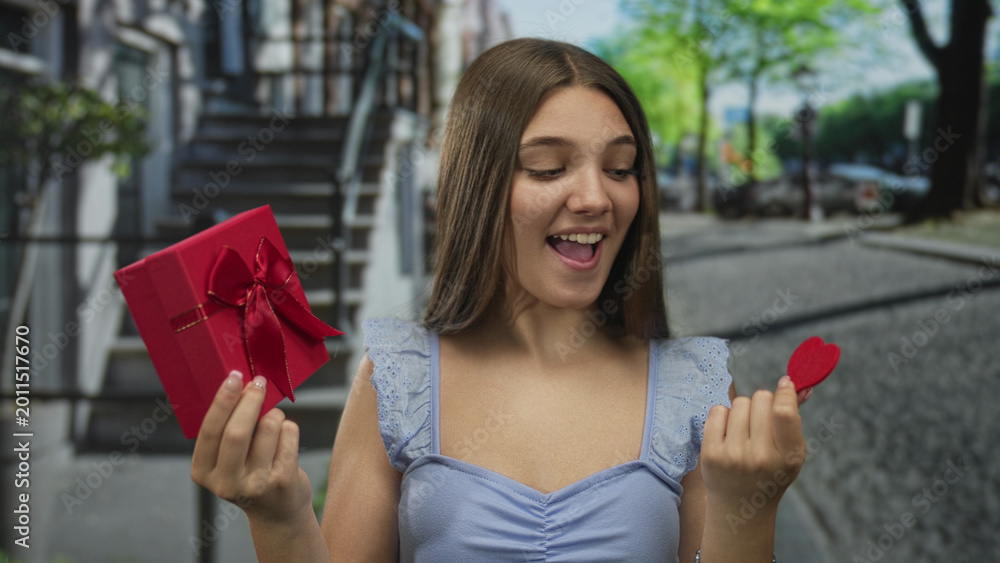Obraz premium Teenage girl holds gift box and heart in hands while smiling on busy urban street under sunlight; romance joy.