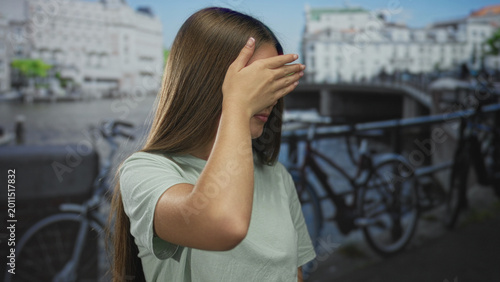 Canvas Print Caucasian girl hand covering eyes gesture on sunny canal street with bicycles and waterway view; shyness