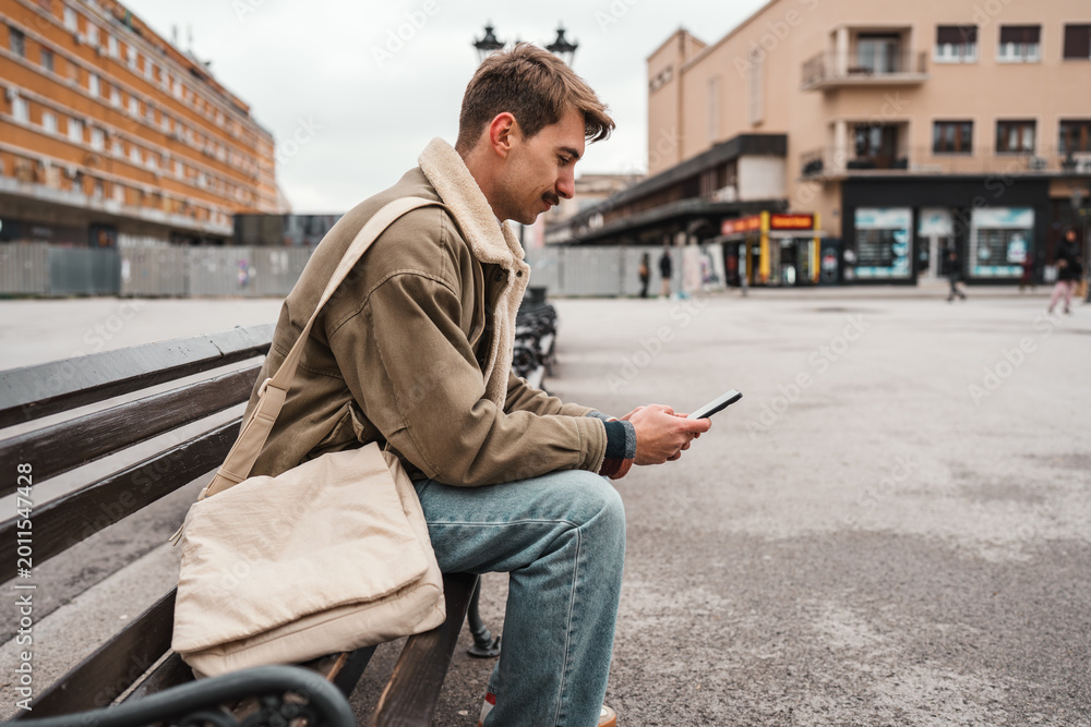 Obraz premium A young man with a mustache and a messenger bag sits on a wooden bench in a public urban square while focused on his mobile phone.