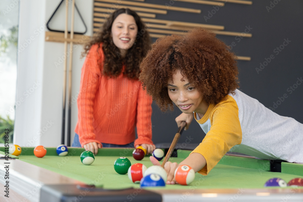 Fototapeta premium Diverse female friends playing at rec room pool table with cue and balls, wearing colorful shirts