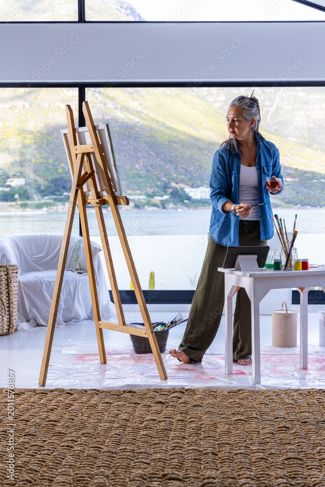 Naklejka premium Senior Asian woman wearing denim standing barefoot at easel in studio painting with brush and glass