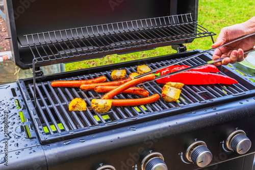Close up view of hand turning sausages on gas grill with roasted vegetables in fresh outdoor setting. Sweden.