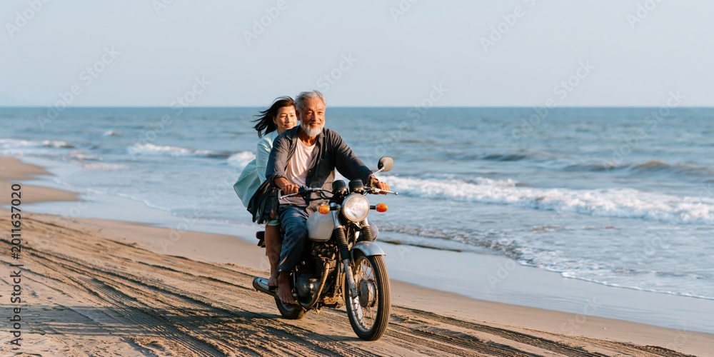 Fototapeta premium Older couple riding motorcycle along shoreline on sandy beach