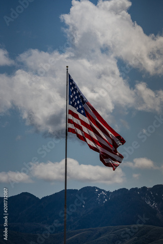 American flag waving in the wind above high  mountains in the distance against blue sky with giant puffy white clouds