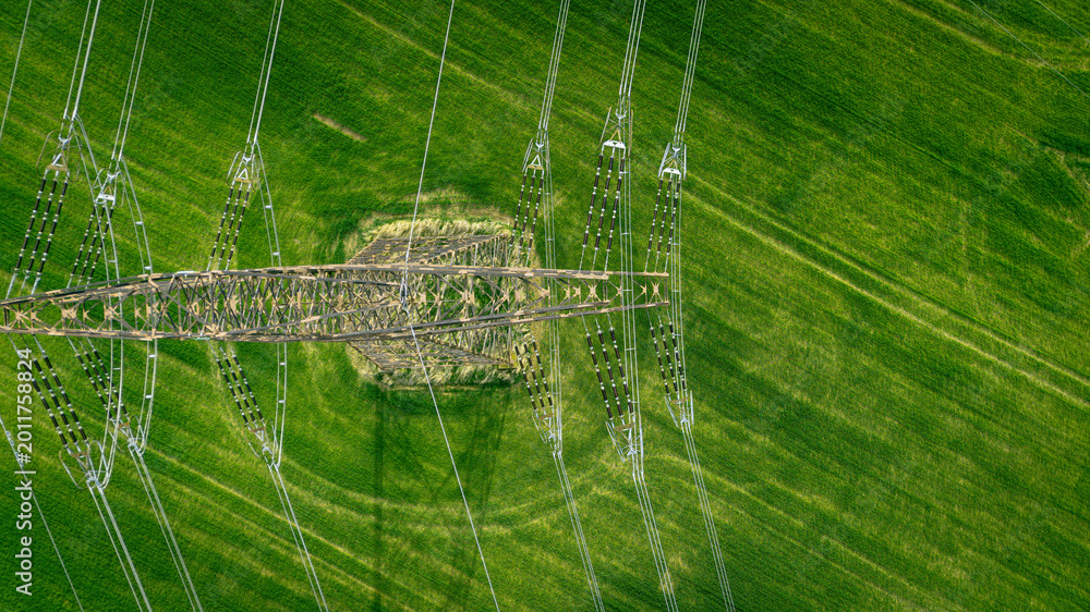 Obraz premium Aerial top-down view of a high-voltage power line pylon in a vibrant green agricultural field. Industrial infrastructure meets nature in a symmetrical composition.