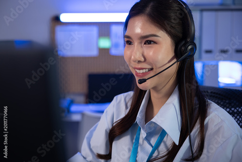 Smiling young asian female customer service representative wearing headset working in office environment showing cheerful emotion assisting clients with friendly confident attitude late at night