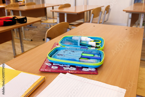 Open pencil case with school supplies on wooden desk in classroom. Pens, pencils, scissors and stationery on table. Back to school concept, elementary education, learning process, student workspace