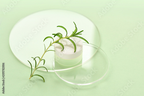 Pastel green background featuring glass transparent discs, cream jar and sprig of rosemary. Natural cosmetics concept