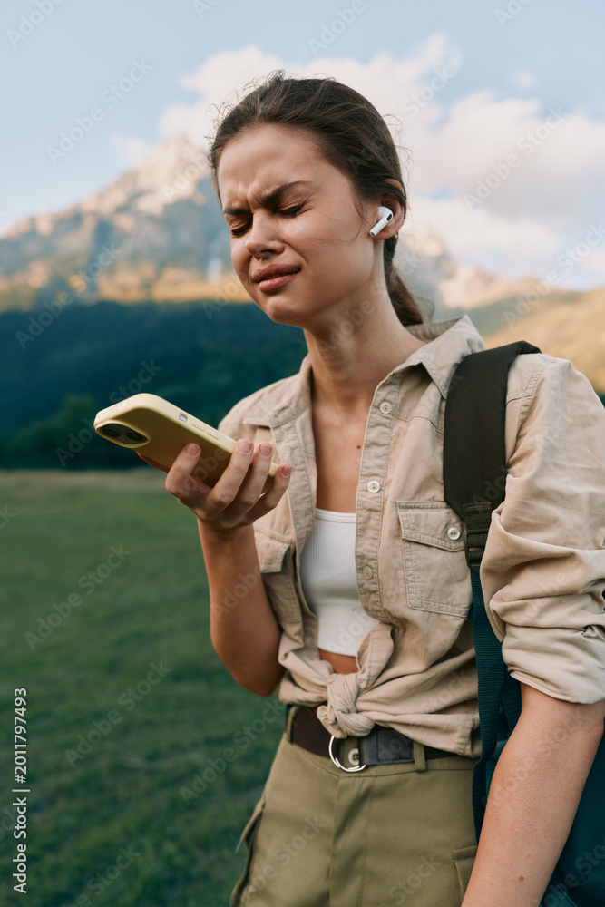 Naklejka premium Smiling woman outdoors checking smartphone during a hike. Casual outfit and backpack against a mountainous landscape. The scene conveys lifestyle, exploration, and wellness.