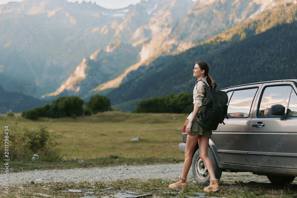 Fototapeta premium Energetic woman outdoors beside a car, exploring mountain scenery, sunlit moment, casual travel gear, serene atmosphere, nature connection, fresh air and freedom in nature under blue sky