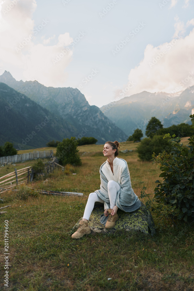 Fototapeta premium Young woman enjoys outdoor lifestyle in a mountain meadow, sits on a rock, wearing light cardigan and boots, tranquil landscape behind, calm mood and natural surroundings