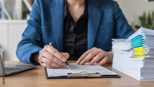 Person in a blue suit signing documents with a stack of papers on the desk paperwork writing