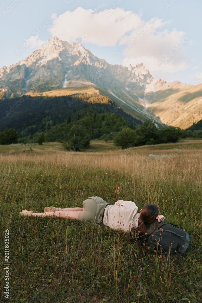 Naklejka premium Calm woman lying in a sunlit meadow with distant mountains, relaxed pose, gentle breeze, natural colors, lush grass, outdoor lifestyle moment soft light, open landscape.