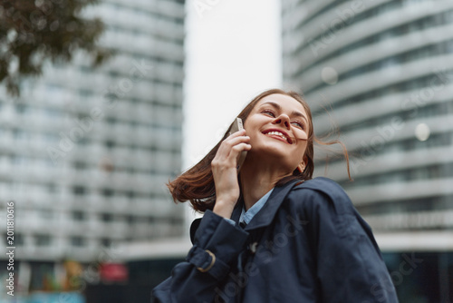 Obraz na plátně Confident woman in a navy jacket smiles while talking on the phone in a dynamic urban setting, tall buildings behind, candid street portrait