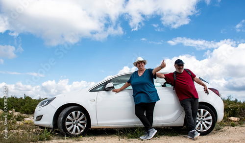 Bild auf Leinwand Pareja de jubilados al lado de su coche en la montaña un día soleado con nubes