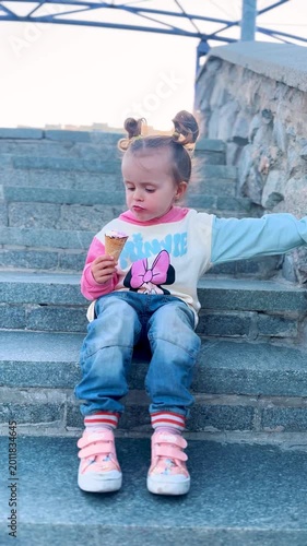 Cute little girl eating ice cream while sitting on stone steps in the city with apartment buildings in the background. Summer lifestyle and happy childhood concept