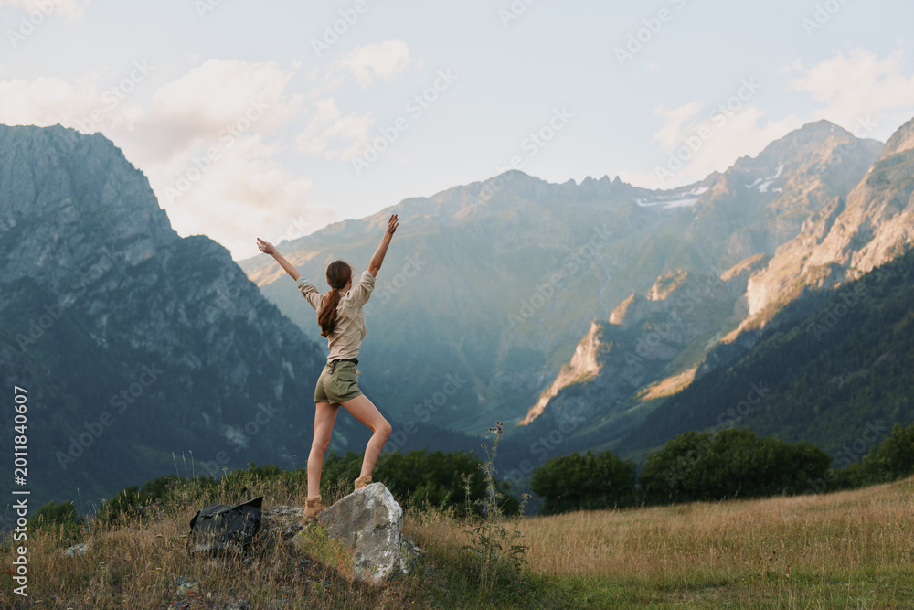 Fototapeta premium Cheerful woman outdoors, standing on a rock with arms raised, mountain valley backdrop, sunny day, carefree mood, nature inspired lifestyle