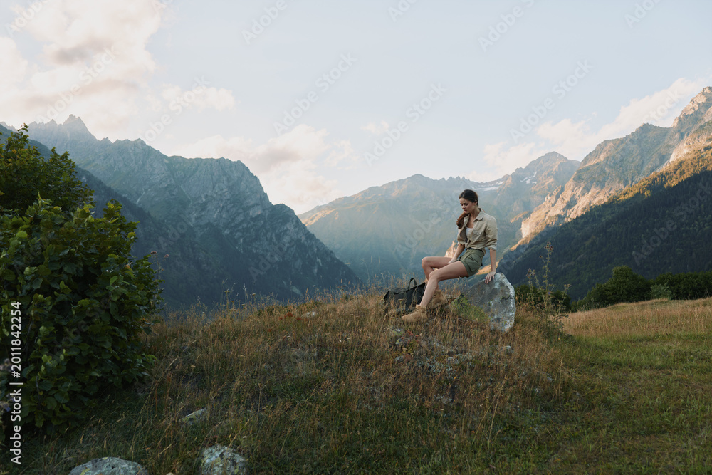 Naklejka premium A woman sits on a rock in a mountain meadow, enjoying the view, calm and reflective. A nature driven moment that conveys outdoor lifestyle and solo exploration.