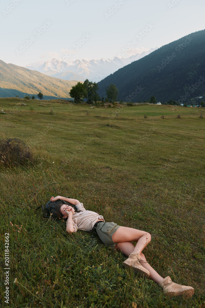 Naklejka premium Cheerful woman lying in a grassy meadow outdoors, sunlit mountains in the background, relaxed pose, gentle breeze, natural colors, carefree lifestyle in bright daylight