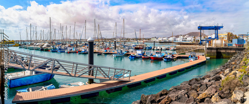 Harbor of Corralejo on Fuerteventura with pier, fishing boats and yachts, turquoise water and coastal town in background under cloudy sky of Canary Islands.