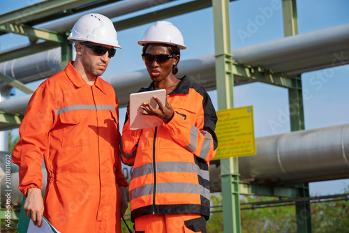 Industrial engineers wearing safety uniforms and helmets inspecting pipeline system at factory, teamwork and maintenance concept, energy industry workers using tablet and documents for technical