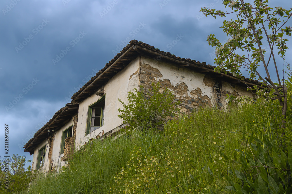 Fototapeta premium Old abandoned rural house on grassy hill under dramatic cloudy sky traditional architecture decay