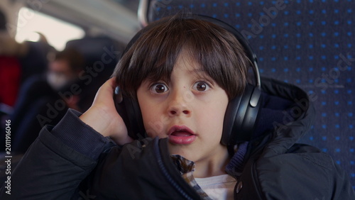 Photography Boy listening with headphones on train seat, reacting with wide eyed surprise an