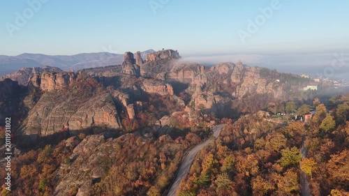 Wide aerial view of a large natural rock massif above autumn forest with low fog drifting across the upper ridge.