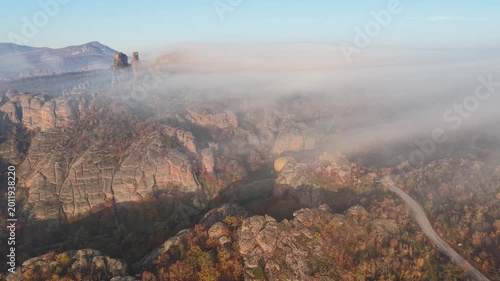 Wide aerial view of a hot air balloon in a rocky autumn landscape with low fog and forest below.