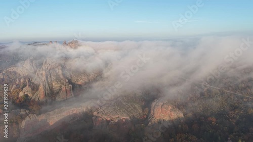 Wide aerial view of natural rock formations partially covered by dense low fog above colorful autumn woodland.