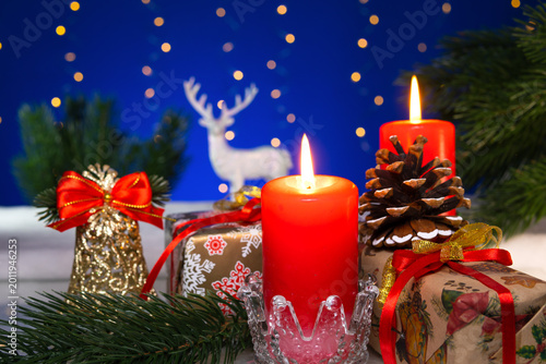 Between two burning candles are gifts with a pine cone and a bell.