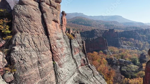 Close aerial pass along natural stone cliffs and a tall rock pillar above colorful woodland.