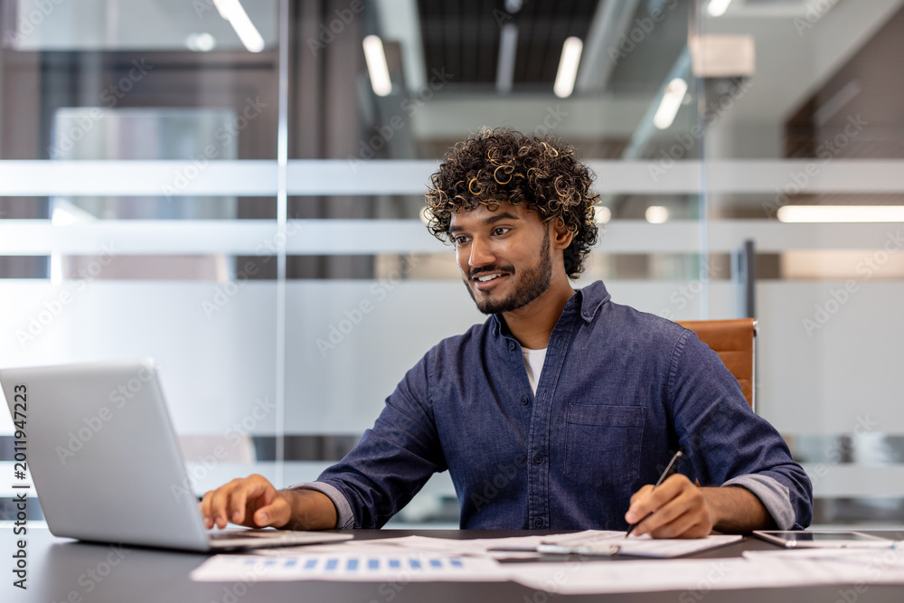 Fototapeta premium Smiling Indian man in the office at the table working with documents and typing on a laptop