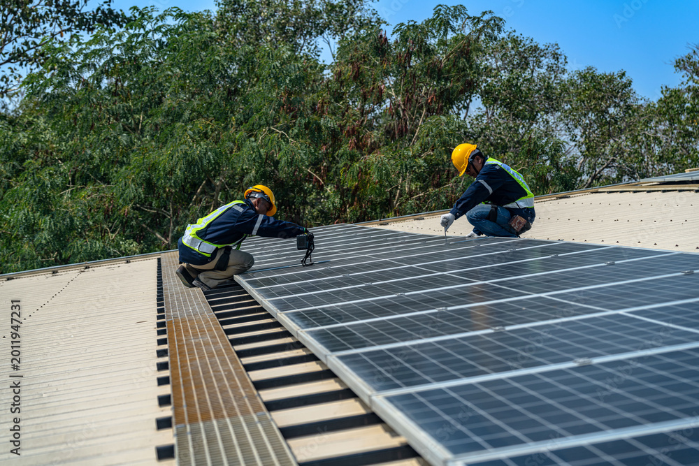 Fototapeta premium A professional technician in safety gear standing on a rooftop, preparing electrical wiring for a solar panel installation under a clear blue sky.