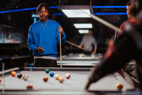 Quadro em tela Young black man holding a cue stick and smartphone, standing by a pool table pla