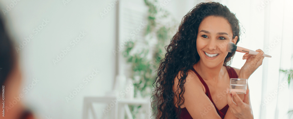 © Prostock-studio - A woman sits in a bright room and smiles as she applies makeup using a brush. She holds a container of powder and looks at her reflection in the mirror.