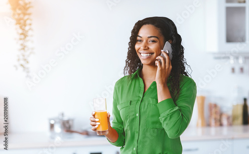 A woman stands in a kitchen while talking on her phone. She holds a glass of orange juice in her hand. The kitchen has a bright and modern look. It is daytime.
