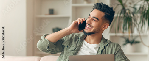 Man is sitting on a couch in a bright room. He is smiling and talking on a phone. A laptop is beside him. There are plants and shelves in the background.