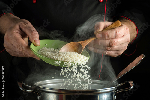 A chef pours rice from a green plate into a pot on the stove. Steam rises from the pot as the chef prepares a meal. The kitchen is busy with cooking