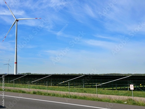 perspective showing a large-scale solar array and wind turbines situated next to a paved road. The dark photovoltaic panels are neatly arranged behind a wire fence, with a tall wind turbine featuring 