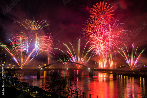 Bild auf Leinwand A sumptuous and dynamic New Year's fireworks display above the illuminated bridge, whose bright red and golden light is beautifully reflected on the surface of the river