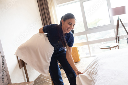 A female hotel staff member is preparing a room for the next guests. She carefully makes the bed, arranges pillows, and ensures the room is tidy and ready for service.