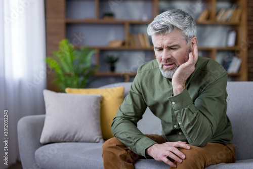 Fotografie Mature man with gray beard sitting on a couch, holding his ear in discomfort, gr