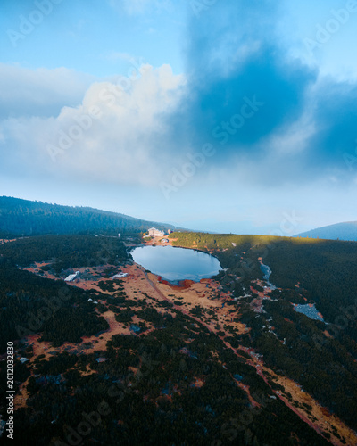 Drone view of a tranquil mountain lake surrounded by dense forest and rugged terrain, with a small lodge or building near the shore. Dramatic clouds and soft light create a moody natural landscape, id