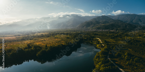 Wide drone panorama of a river or lake winding through green countryside with a small road leading into the distance and mountains under soft atmospheric light.