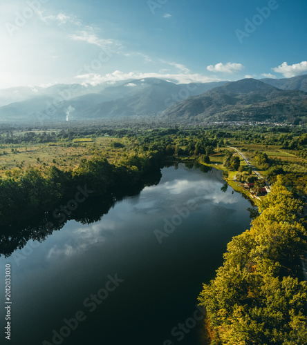 Drone view of a calm lake surrounded by dense green forest and open countryside with mountains in the background. Soft golden hour light and reflections create a peaceful natural scene.