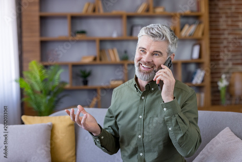 Happy mature man using a smartphone for a conversation, expressing joy and confidence while sitting comfortably at home and engaging in modern communication