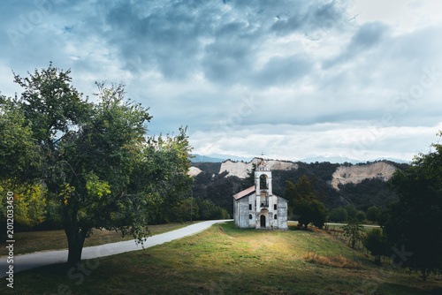 фотографія Melnik, Bulgaria - 09 26 2026: Small old rural church standing alone in a grassy field surrounded by trees and mountains under a dramatic cloudy sky