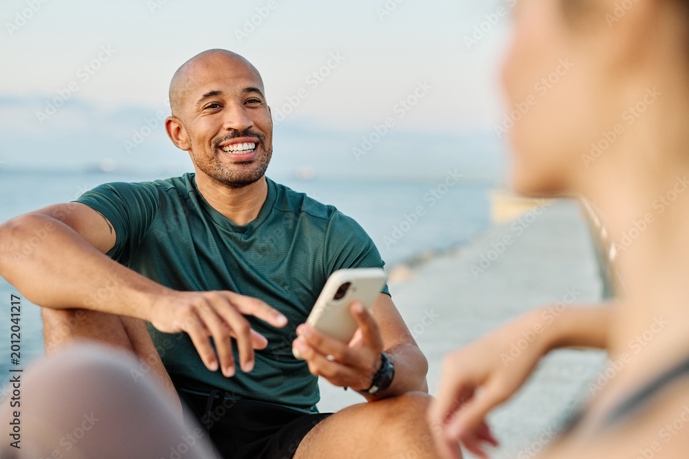 Obraz premium Smiling active young couple jogging exercising and having fun and laughing together taking a breakand using a mobile phone after walking and running in the park, fitness and healhty living concept