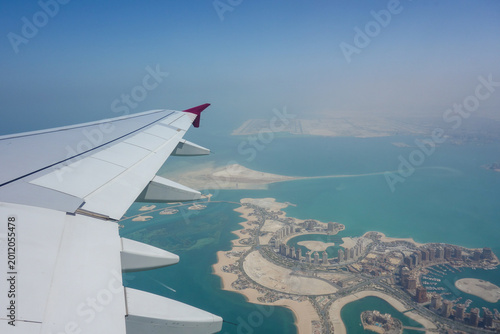Aerial view from airplane window over The Pearl-Qatar artificial island in Doha, Qatar, with aircraft wing visible above turquoise waters, luxury marinas and modern waterfront architecture during flig